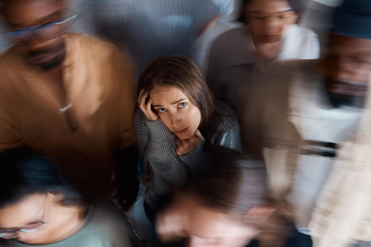 A woman in a gray sweater resting her hand against her head, looking distressed among a blurred crowd