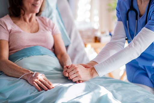 woman laying in hospital bed cared for by nurse holding her hand.